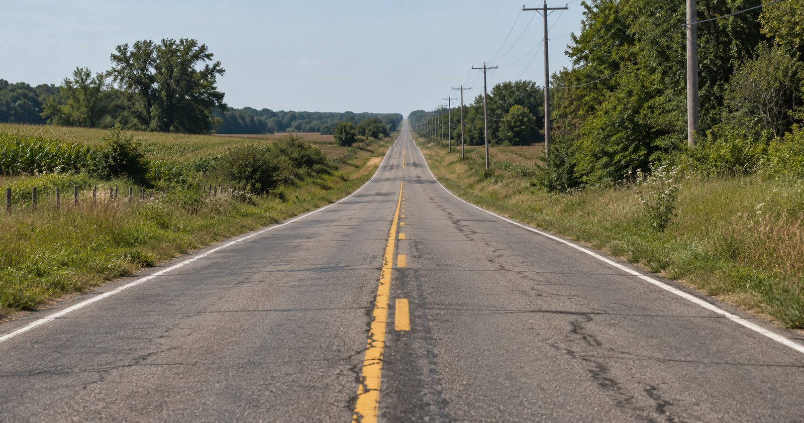 A long, straight rural two-lane road with no shoulder between fields and trees.