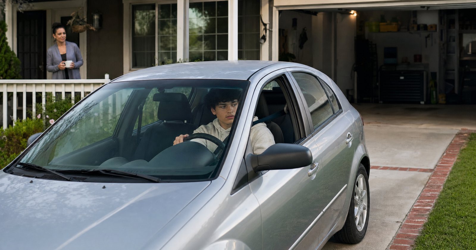 A mother watches from the porch as her teenage son backs out of the driveway alone.